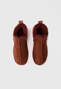 Chaussons en daim brun avec un intérieur moelleux. Ils présentent une couture sur le devant et une petite languette pour un enfilage facile.