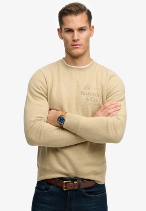 Man met gekruiste armen, gekleed in een beige trui met het Superdry & Co-logo, blauwe jeans, een bruine riem en een horloge met een blauwe wijzerplaat en een kastanjebruine leren band.
