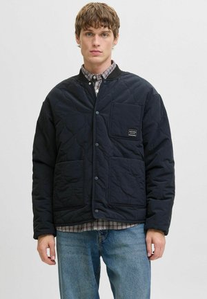 Veste matelassée bleu marine avec fermeture à boutons, deux poches avant et un patch d'étiquette, portée sur une chemise à carreaux et un jean en denim.