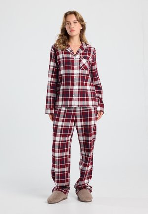 Femme debout portant un ensemble de pyjama à carreaux rouge, blanc et gris avec poche, associé à des chaussons en peluche beige, sur fond blanc uni.
