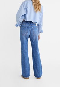 High-waisted flared jeans in medium blue denim with a faded finish, two back pockets, and a raw hem. Paired with a light blue striped crop shirt.