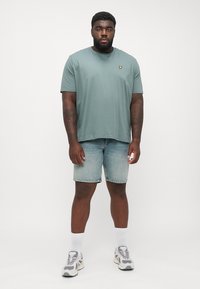 T-shirt verde chiaro a maniche corte con un piccolo logo giallo, abbinata a pantaloni corti di denim sbiadito e calzini bianchi, completata da sneakers grigie e bianche.