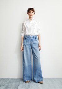 Helles blaugraues weites Beinjeans mit hoher Taille, kiefendesign mit fünf Taschen, sichtbaren Nähten und einem leichten Verblassen. Kombiniert mit einem weißen Hemd.