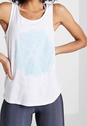 Ärmelloses weißes Tanktop mit hellblauem geometrischem Blumenmuster, getragen zu dunkel gestreiften Leggings, Hände in die Hüften gestützt.