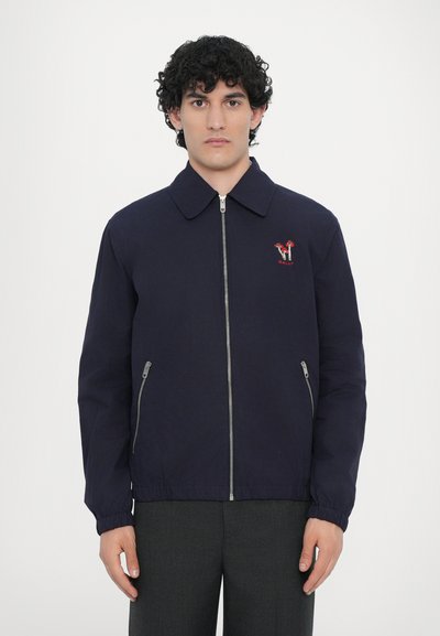 Giacca blu navy con colletto, zip frontale, tasche laterali e dettaglio del logo ricamato rosso. Il materiale è liscio con una finitura leggermente lucida.