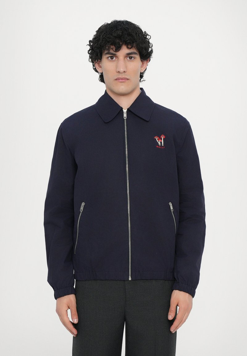 Giacca blu navy con colletto, zip frontale, tasche laterali e dettaglio del logo ricamato rosso. Il materiale è liscio con una finitura leggermente lucida.