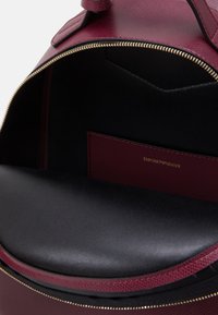 Interiör i vinrött läderryggsäck med en slät svart foder, guldfärgat blixtlåsförslutning och en märkt vinröd ficka med präglad text.