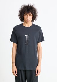 Juodas medvilnės marškinėlis su apvalia iškirpte, ant priekinės pusės esantis baltas Nike logotipas ir geometriniai raštai, kurie yra įvairių formų ir dydžių.