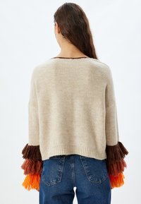Maglione beige con maniche oversized e frange marroni, arancioni e arancioni vivaci. Orlo rifinito e vestibilità rilassata. Indossato con jeans blu.