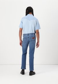 Camicia di jeans azzurro chiaro con maniche corte, abbinata a jeans a gamba dritta blu medio. Scarpe nere con texture liscia. Accento in pelle marrone sui jeans.