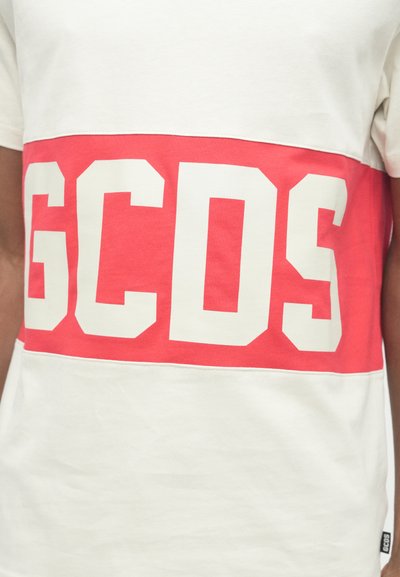 T-shirt bianco con una ampia striscia rossa e grande lettering bianco "GCDS", in tessuto di cotone, scollo rotondo, maniche corte e dettaglio con etichetta laterale.