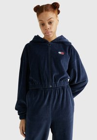 Marineblauwe velours hoodie met rits, elastische taille, capuchon en logo patch. Zachte textuur; relaxte pasvorm met manchetten en zakken.