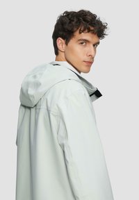Veste légère grise, imperméable, avec capuche, dotée d'une fermeture éclair et d'accents noirs. Sa texture douce et sa coupe décontractée offrent confort et mobilité.