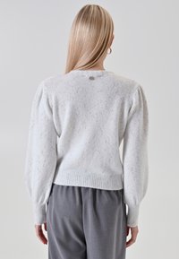 Donna con lunghi capelli biondi che indossa un maglione grigio chiaro a pois con maniche a palloncino e pantaloni grigio scuro, in piedi davanti a uno sfondo semplice.