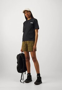 Camiseta negra de manga corta con bolsillo en el pecho, combinada con pantalones cortos cargo de color verde oliva. La mochila negra y las botas hasta el tobillo completan el conjunto.