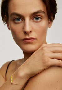 Pulsera de esmalte amarillo con cadena de oro, con detalles grabados, llevada en una muñeca desnuda contra un fondo neutral.