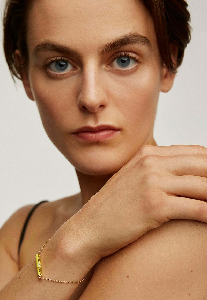 Pulsera de esmalte amarillo con cadena de oro, con detalles grabados, llevada en una muñeca desnuda contra un fondo neutral.