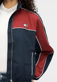 Μπουφάν με μπλοκ χρωμάτων σε σκούρο μπλε και κόκκινο, με φερμουάρ στο μπροστινό μέρος, λευκή επένδυση και έμβλημα με λογότυπο. Κατασκευασμένο από ελαφρύ υλικό με λείο υφή.