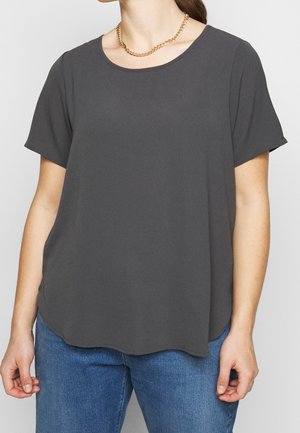 Kvinde iført en mørkegrå kortærmet skjorte, blå jeans og en guldkæde mod en ensfarvet baggrund.