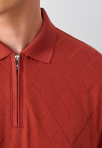 Polo en tricot rouge avec un col zippé, texture en motif diamant et un design de col uni.