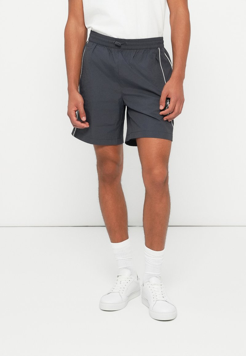 Dark gray shorts with elastic waistband, side pockets, and white piping details. Worn with white socks and sneakers.