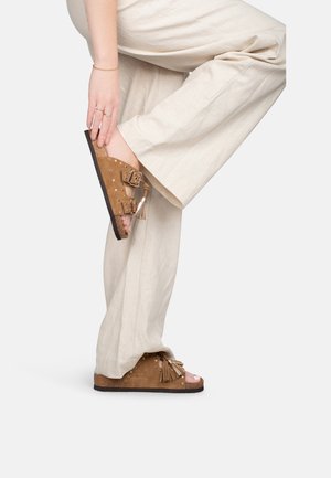 Personne portant un pantalon beige à jambes larges et des sandales compensées brunes avec des pampilles et des clous métalliques, levant un pied pour toucher le talon de la sandale.