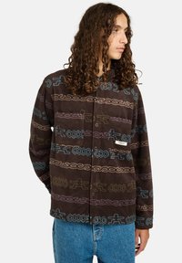 Persona con lunghi capelli ricci che indossa una giacca in pile marrone a fantasia con disegni blu e viola e jeans blu, che guarda di lato.