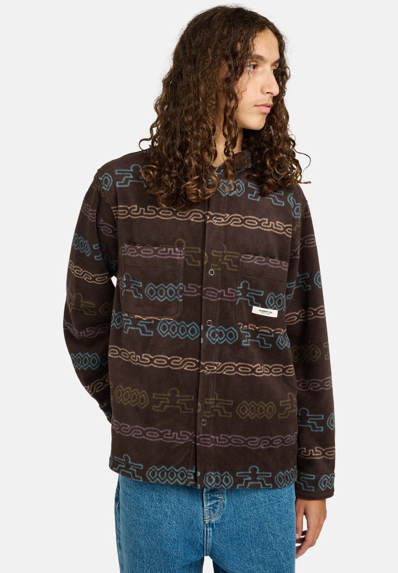 Persona con lunghi capelli ricci che indossa una giacca in pile marrone a fantasia con disegni blu e viola e jeans blu, che guarda di lato.