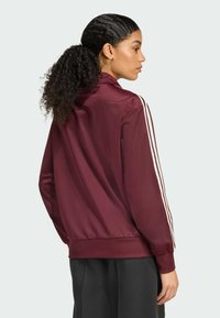 Maroon athletic jacket with white stripes along the sleeves, smooth texture, zip up collar, and fitted waistband. Back view shown.