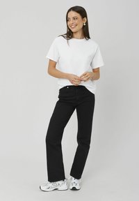 T-shirt en coton blanc associé à un jean noir taille haute. Le modèle porte des baskets blanches et des boucles d'oreilles créoles, avec un fond neutre.