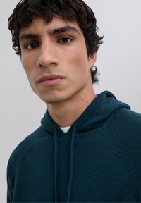 Ung man med mörkt lockigt hår som bär en teal hoodie, ser direkt in i kameran med ett neutralt uttryck mot en enfärgad bakgrund.