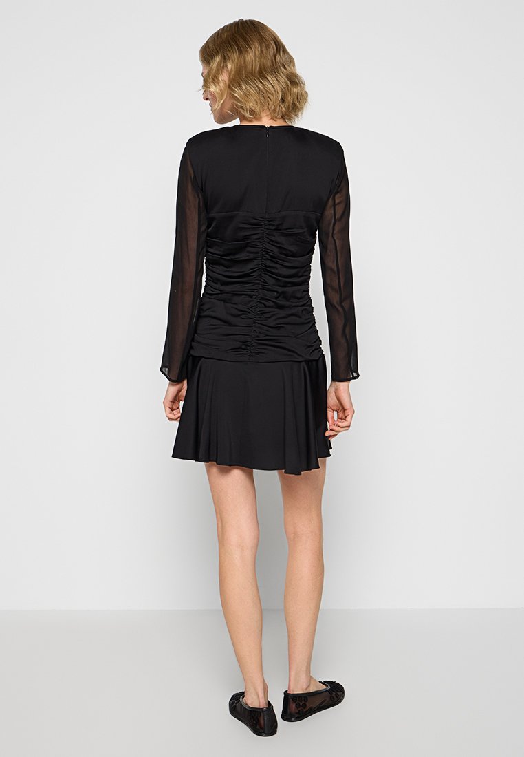 Vestido negro con mangas largas transparentes, detalles fruncidos en la parte posterior y una falda acampanada. Lleva zapatos negros tipo mocasín.
