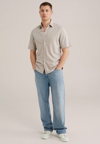 Cămășă cu mânecă scurtă, bej deschis, cu un model geometric, purtată cu blugi deschiși la culoare, largi, și adidași albi.