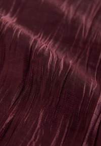 Tissu texturé bordeaux avec des motifs ondulés verticaux, un éclat lisse et une apparence soyeuse, suggérant un matériau léger et facile à draper.