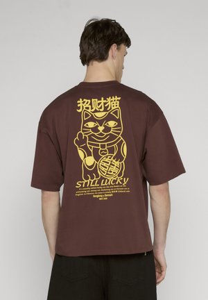 Mand iført brun t-shirt med stort gult motiv af en vinkende heldig kat og teksten "STADIG HELDIG" på bagsiden.