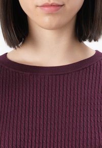 Femme aux cheveux courts et foncés portant un pull bordeaux texturé, montrant le bas du visage, le cou et les épaules sur un fond blanc.