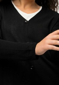 Schwarze Strickjacke mit V-Ausschnitt, langen Ärmeln und einem einzelnen schwarzen Knopfverschluss. Getragen über einem weißen Oberteil.