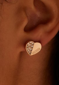 Boucle d'oreille en forme de cœur en or rose avec une surface texturée. Un côté est orné de strass, l'autre présente le nom de marque gravé "GUESS."