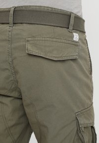 Pantaloni cargo verde oliva realizzati in cotone misto, con una cintura testurizzata e una tasca posteriore con pattina. Piccola etichetta con logo visibile sulla vita.