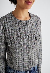 Veste courte à carreaux gris et multicolores avec des manches longues, dotée de deux poches poitrine et de quatre boutons. Tissu texturé.