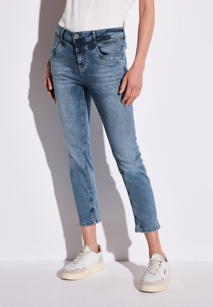 Jean en denim bleu clair, coupe slim, avec cinq poches et une fermeture à boutons. Portés avec des baskets blanches et beiges, présentant des accents texturés.