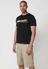 Camiseta de algodón negra con texto gráfico en amarillo y gris; combinada con pantalones cortos de cargo beige que tienen bolsillos laterales y una pequeña etiqueta.