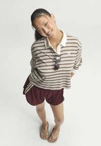 Ung kvinde smilende og blinker, iført en stribet poloshirt, bordeaux shorts, klipklappere og solbriller hængende fra hendes skjorte.