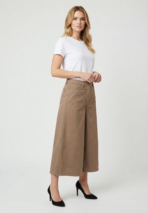 Femme debout portant une chemise blanche à manches courtes, un pantalon beige taille haute, large et raccourci, et des chaussures à talons noirs sur fond blanc.