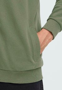 Grüner Sweatshirt mit weicher Textur, gerippten Bündchen und einer Seitentasche. Der Saum ist in einem ähnlichen gerippten Muster für zusätzlichen Komfort verarbeitet.