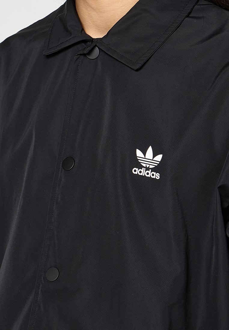 Nahaufnahme einer Person, die eine schwarze Adidas-Jacke mit Kragen und schwarzen Druckknöpfen trägt, auf der sich ein weißes Adidas-Logo auf der Brust befindet.