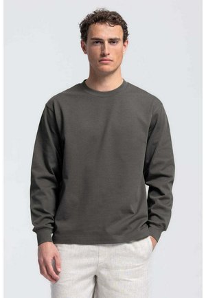 Man met krullend haar, gekleed in een donkergrijs shirt met lange mouwen en een lichte broek, staand tegen een effen lichte achtergrond.