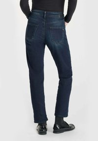 Jean en denim bleu foncé avec une coupe droite, un design à cinq poches et une finition texturée, associés à des chaussures noires brillantes et des chaussettes.