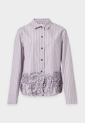Femme portant une chemise rayée violet et blanc à manches longues avec des détails floraux, associée à un pantalon blanc à jambes larges et des ballerines noires pointues.