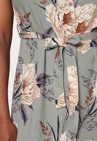 Kukkaldressi vihreästä kankaasta, jossa on solmittava vyötärö, suuria beige-kukkia, tummia lehtiä ja nappeja edessä. Sileä rakenne.